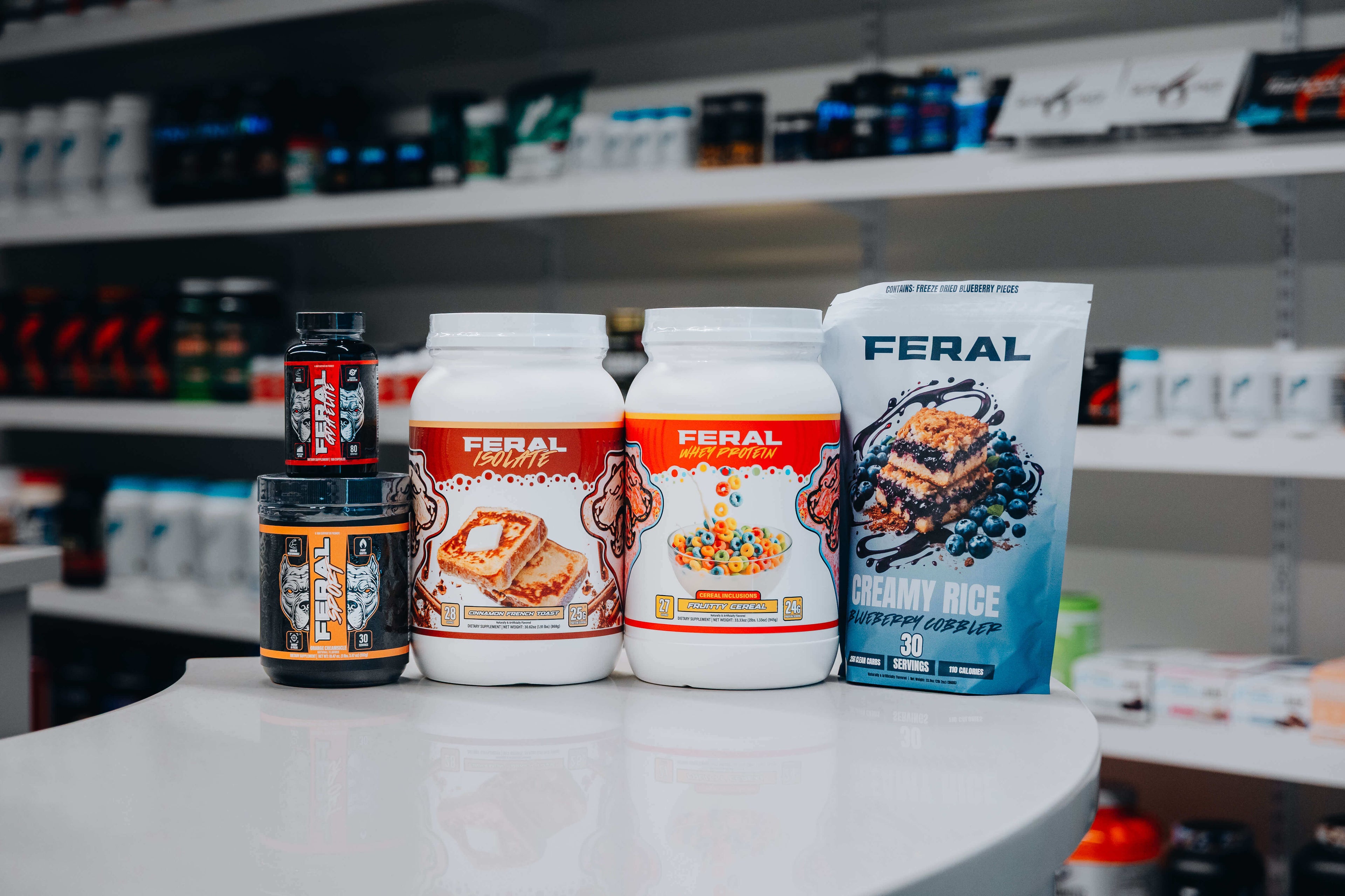Five Feral brand supplements, including protein powders, pre-workout, and creamy rice, arranged on a white counter inside a supplement store.